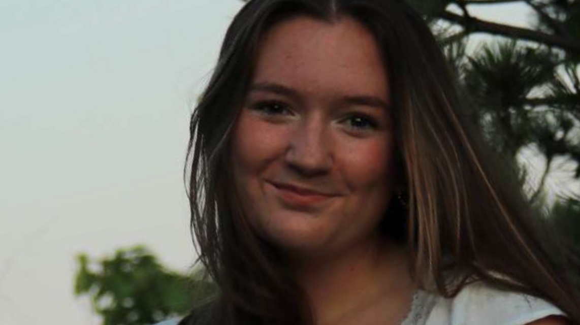 A young woman with long brown hair smiling slightly, wearing a light-colored top, with trees and sky in the background.